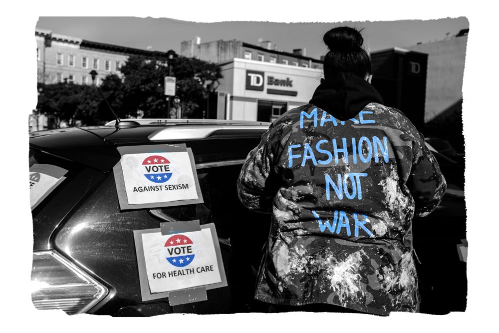 A woman wearing a camouflage jacket is standing near her car with her back turned to the camera. On the back of her jacket are the words, "Make Fashion Not War." Taped to the car are signs encouraging voting.