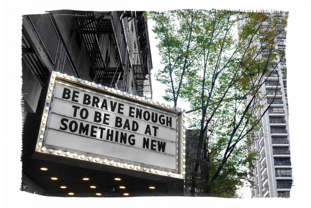 A rectangular billboard with small, lit, white lightbulbs around its border is mounted on a city building below a fire escape. The bottom of a skyscraper style building can be seen in the background down the block. Black capital letters on the sign are arranged to say, "Be Brave Enough To Be Bad at Something New."