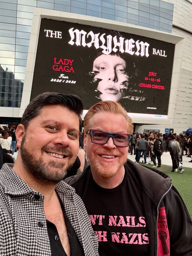 One bearded Brunette man and one bearded Ginger man standing in front of SF’s Chase Center and a billboard with Lady Gaga: The Mayhem Tour image.
