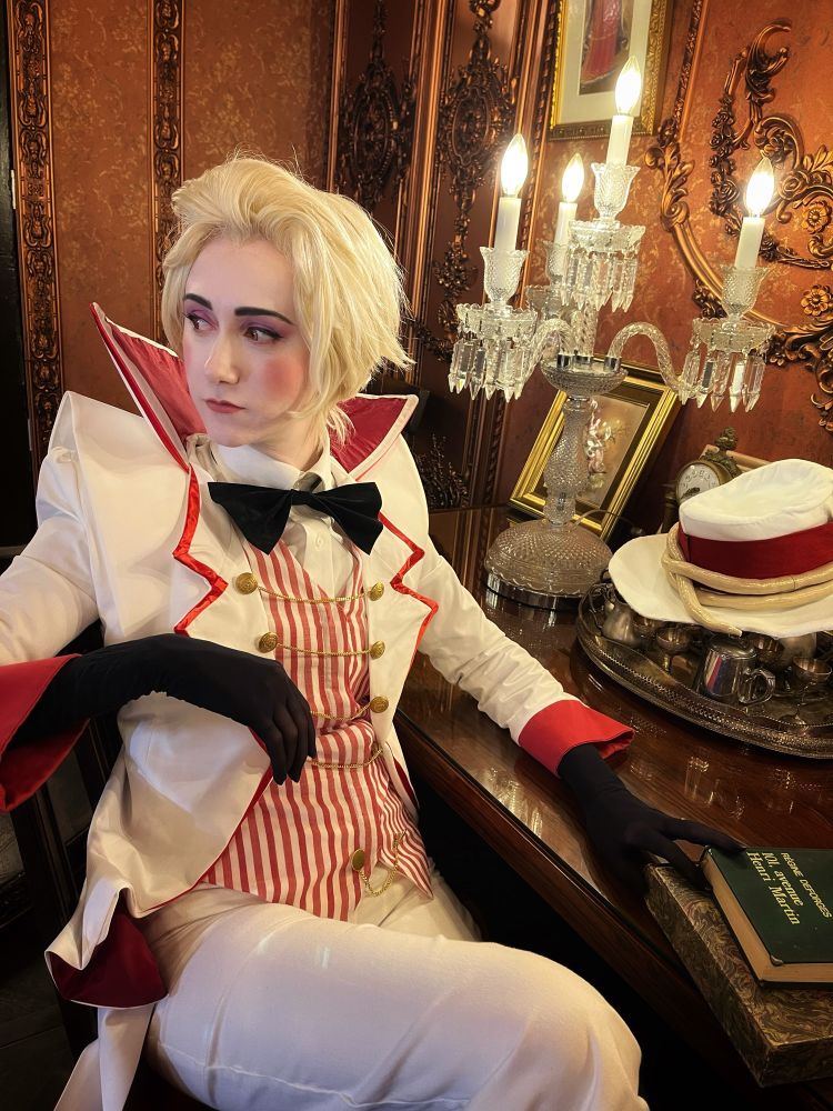 Kei sits at a fake-candle lit desk in Lucifer Morningstar cosplay from
Hazbin Hotel. Lucifer’s hat is removed and he looks moody about human’s choices. 