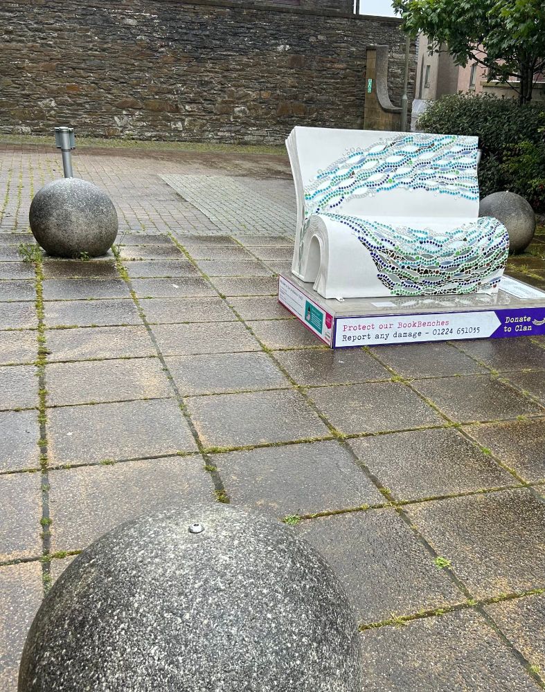 Courtyard in front of Orkney library. Paving stones on the ground, three stone balls in view and also a bench in the shape of an open book with a shoal of mosaic fish swimming across its back and seat