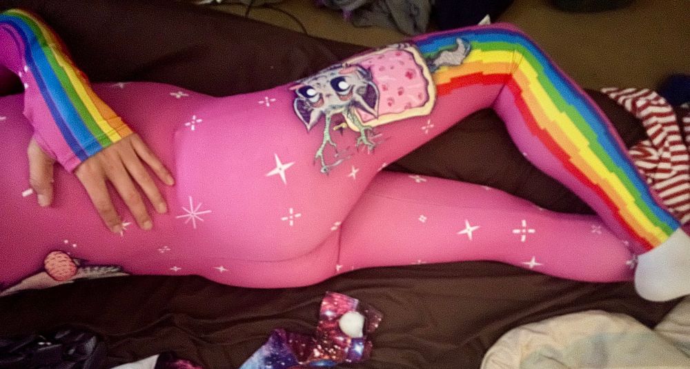 Wearing a pink “fit onesie” from Badinka, a hot pink spandex catsuit with a Nyan-cat inspired character and rainbows down the sleeves and legs. 
Side view, reclined on bed. Showing a lil booty.