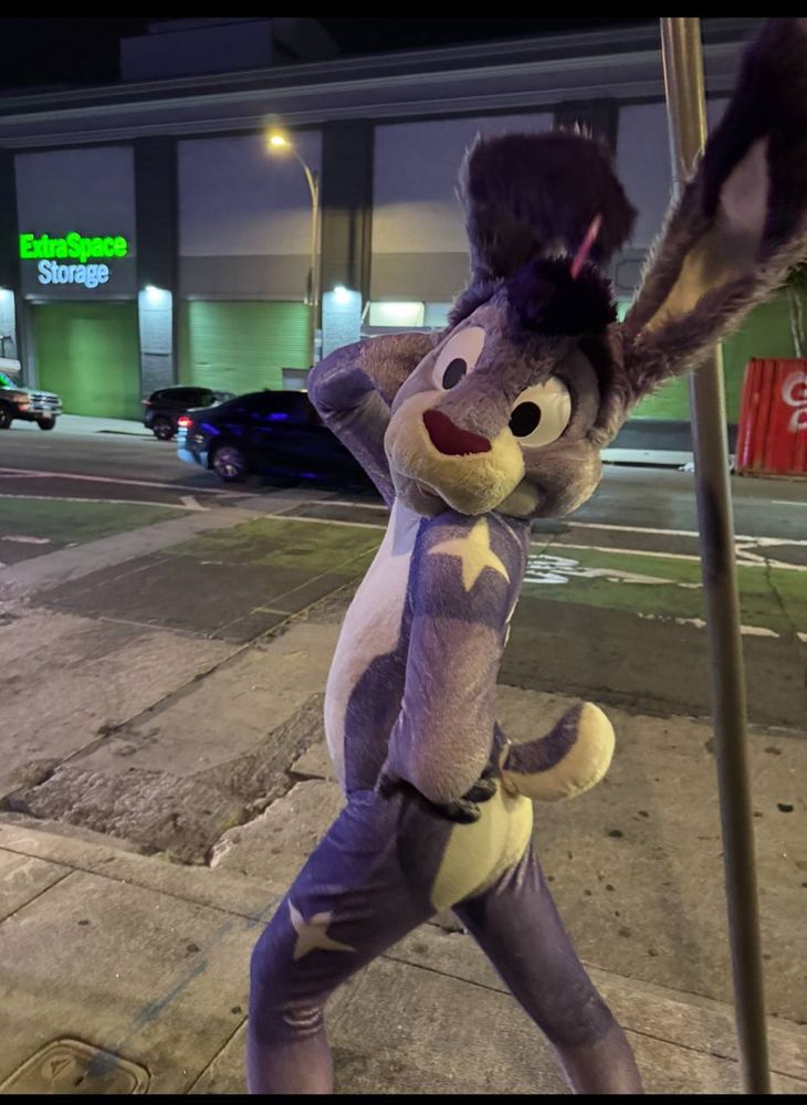Twilight in Fur Petsuit by ForFun Bangkok, purple bunny posing in his signature pose, side view twisting left to face the camera, one paw on hip, other paw behind head. Outside Foundry in San Francisco at the semi monthly Frolic party
