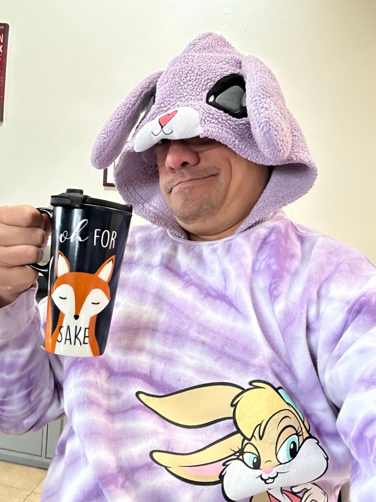 Jimmy (the human behind Twilight bunny and Yippee Coyote) grinning in a purple bunny sleeper with the hood pulled over his own eyes, showing bunny eyes and ears. Jimmy is wearing a tie dyed light purple Lola Bunny sweatshirt and is holding a coffee mug reading “oh for 🦊 sake”