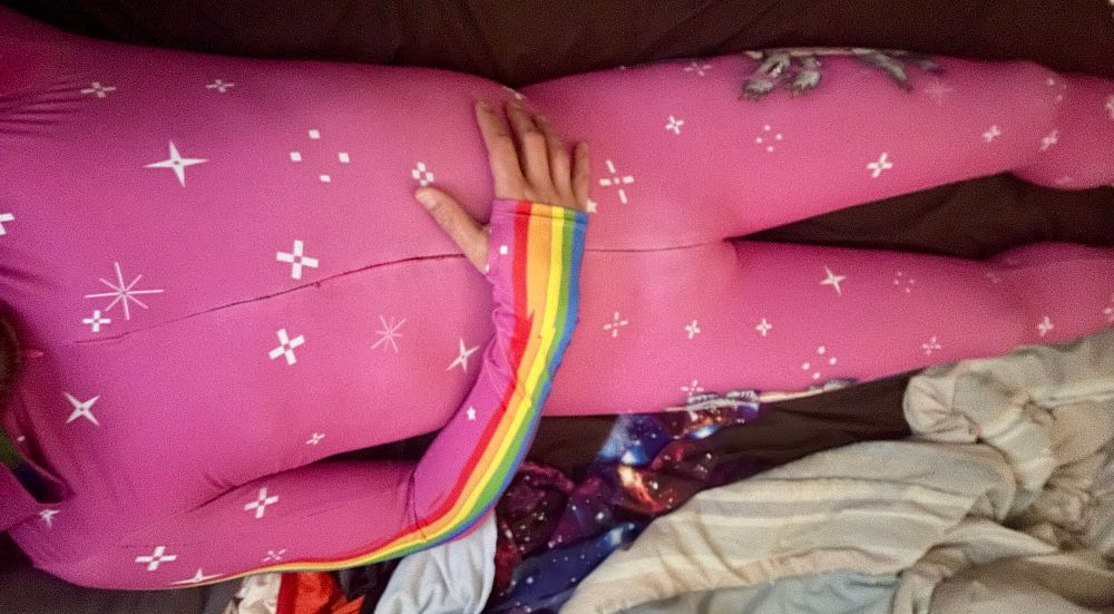 Wearing a pink “fit onesie” from Badinka, a hot pink spandex catsuit with a Nyan-cat inspired character and rainbows down the sleeves and legs. 
Front view, reclined on bed. Front is mostly in decorated.