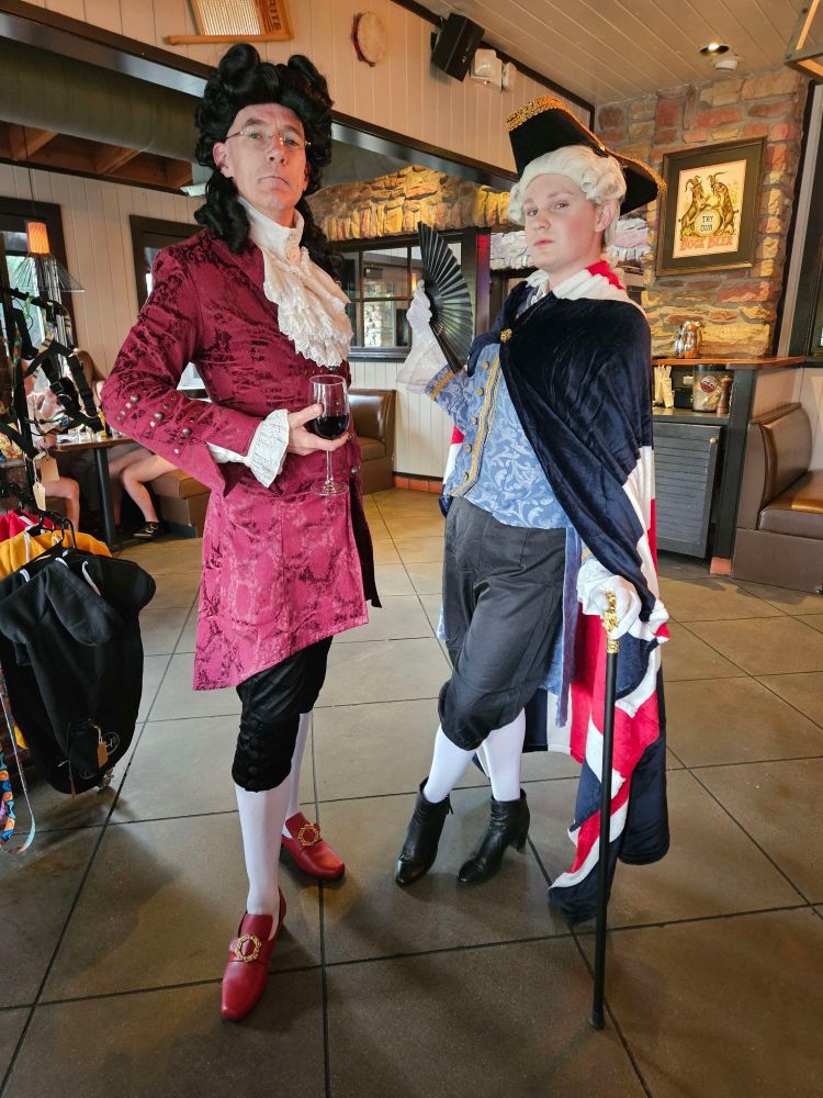 A pair of 18th century attired gentlemen, one in a red velvet coat and lace, with high heeled red shoes, with a black ringlets wig, holding a glass of red wine, the other in a blue brocaded tunic and darker blue Union Jack cape, with a black fan in one hand leaning on a cane, wearing a white ponytailed wig and a black tricone hat, and a hint of blush on his cheeks and black high heeled boots.

Both are in white tights, naturellement.