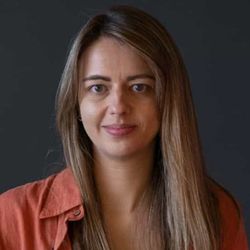 A young woman with pale skin, long straight light brown hair and a peach shirt looks smilingly into the camera. There is a dark background behind her.
