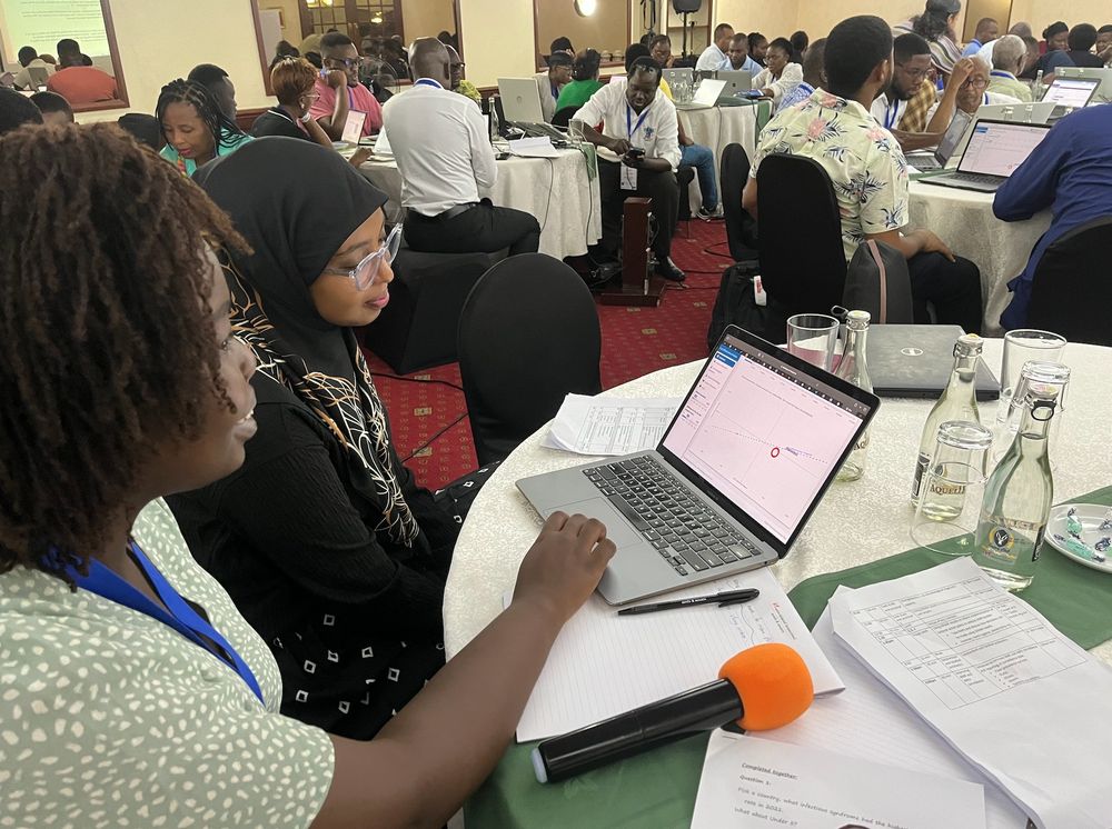 Rachel Otuko (@missotuko.bsky.social), on the left of the picture, an Oxford DPhil student and teacher on the workshop explains a graph plotted on the laptop screen to a student from the Africa CDC attending the workshop. The room is filled with other students who are deep in thought while staring at computer screens which are running the same R Shiny Application.
