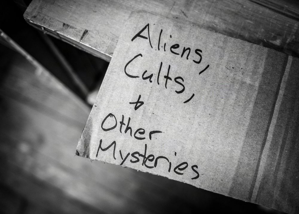 A black and white photo looking down onto a  simple handmade cardboard sign with the words "Aliens, Cults & Other Mysteries" written on it with black felt pen. 