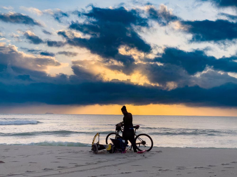 A color photo taken at sunrise on Miami Beach. A silhouette of a man standing next to his bicycle and small bicycle trailer. In the background is the Atlantic Ocean and a beautiful sunrise with the sun behind a band of dark clouds. 