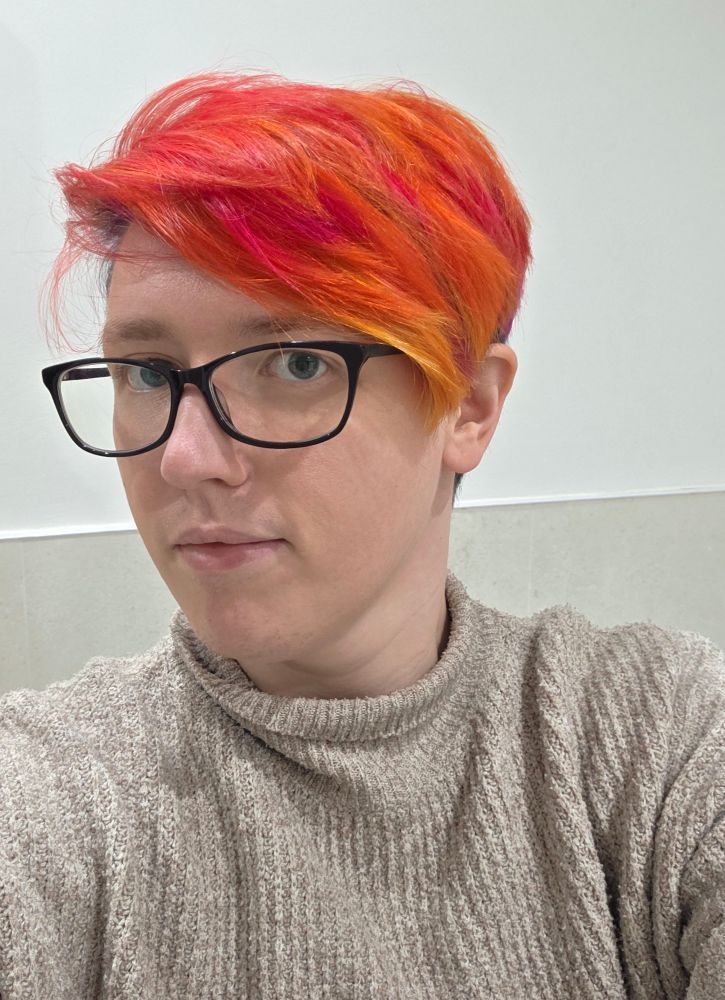A photo of a nonbinary person with short bright red, orange and yellow hair. They are wearing large black glasses and a grey turtleneck jumper.