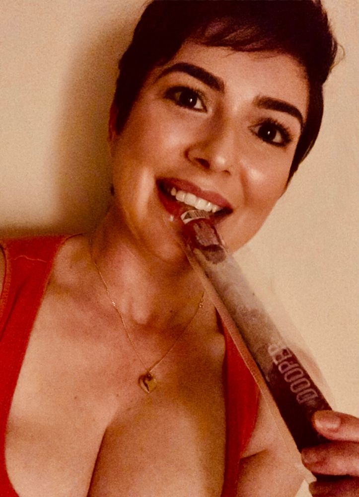 A woman with short brown hair, wearing an orange singlet is smiling while enjoying a purple zooper dooper