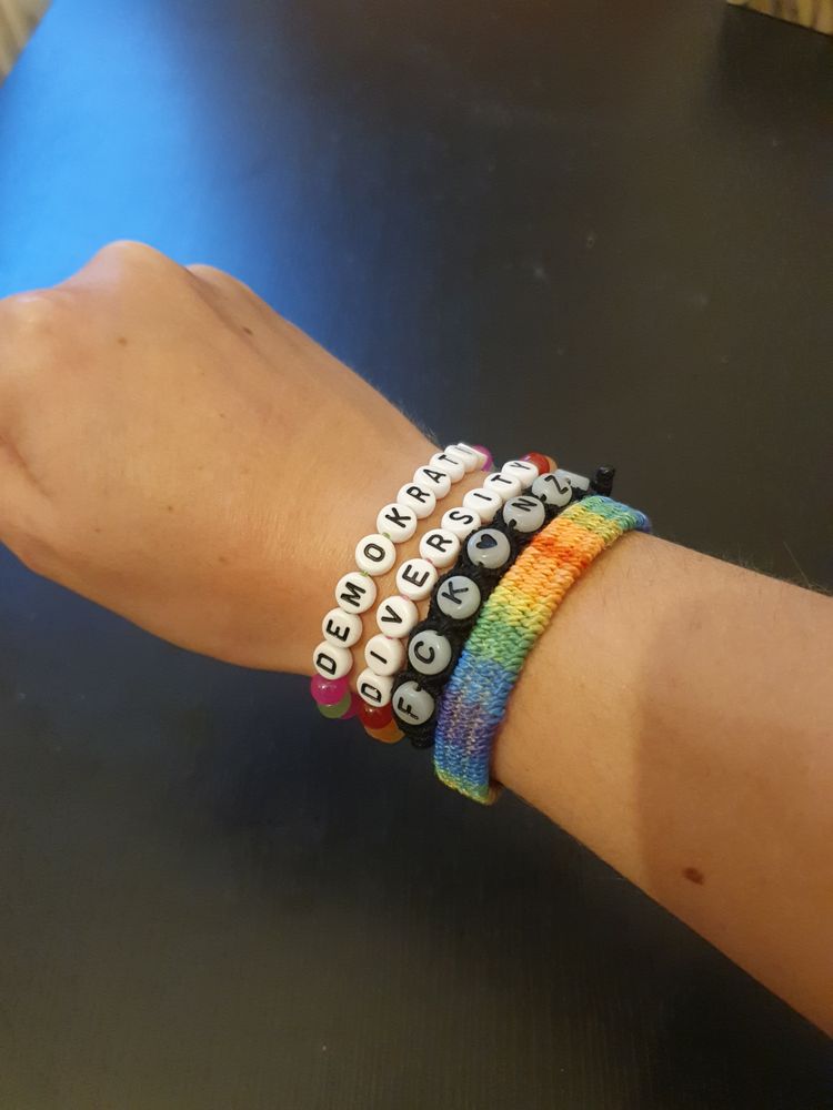 Foto von meiner zur Faust geballten Hand mit vier Armbändern am Handgelenk:
Zwei alte (ein geknüpftes Band in Regenbogenfarben und ein schwarzes Band mit den Buchstaben FCK NZS), und zwei neue Perlenarmbänder. Das erste hat die Buchstaben für Diversity, das zweite für Demokratie.