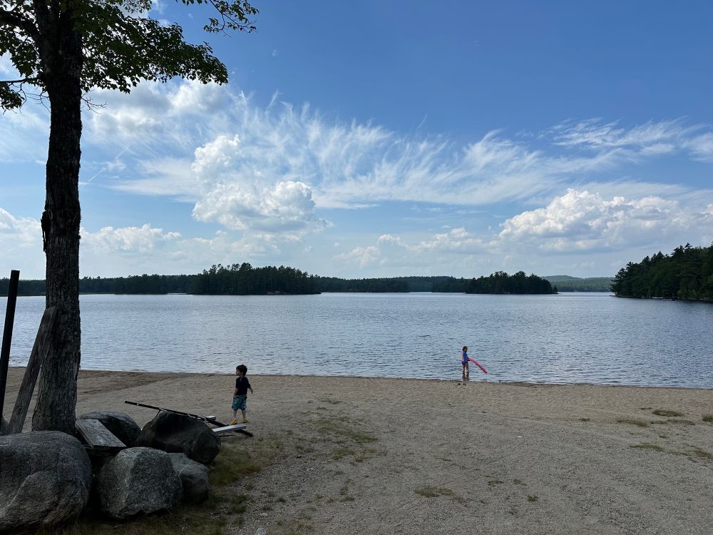 One of Maine’s few sand beaches, empty except for a tree, pile of rocks, and 2 kids. There are a few islands covered in evergreens. The water is calm (as far as we can tell), the sky a perfect blue with both wispy and fluffy clouds