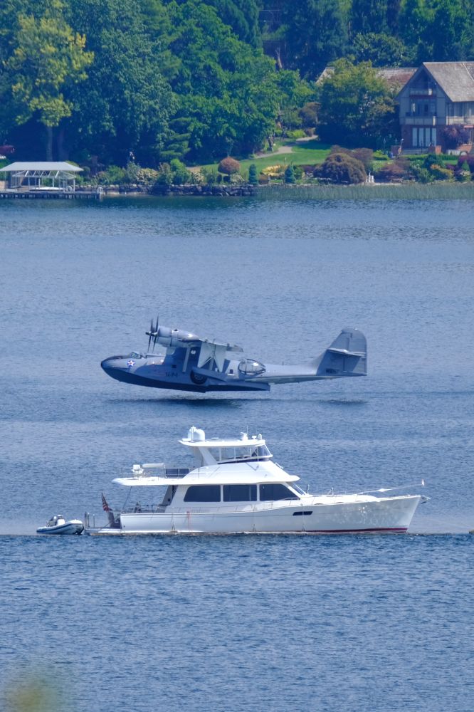 A gray Catalina PBY comes in for landing by a white cabin cruise on a lake 