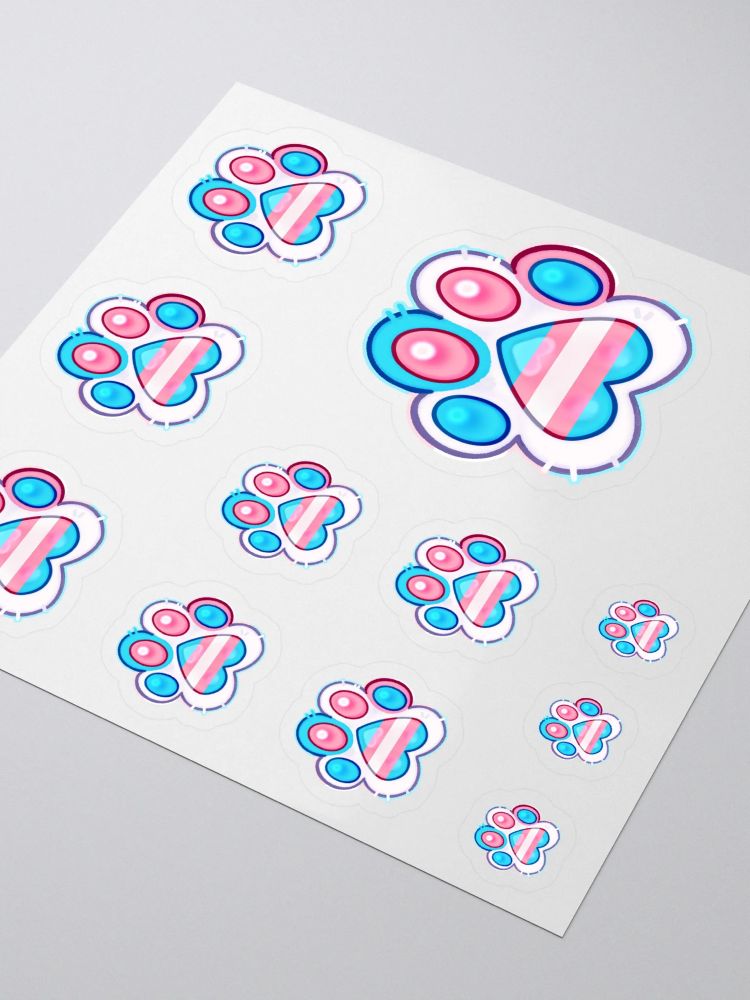 A digitally drawn sticker design of a paw with the Transgender Flag colours. The middle paw pad is shaped like an upside down heart, with the Trans flag stripes inside of it. 

The majority of the paw is white, with one blue toe and one pink toe. The paw pads (toe beans!) alternate between blue and pink. Little tufts of fur stick out. A thin outline surrounds it, in a much lighter version of the transgender flag colours.

A mockup product photo of the above paw design with the trans flag colours, on a sticker sheet. The stickers variety in size.

The sticker sheet lays on a grey surface. 