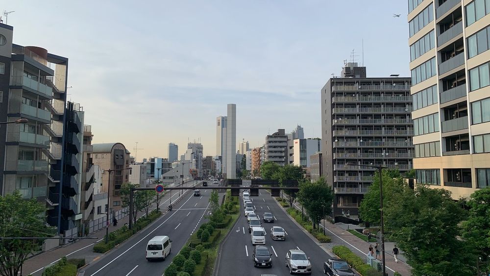This photo was taken from the pedestrian bridge at Tomigaya intersection in Shibuya Ward, looking north toward the road. The road slopes gently upward, with two towers standing in the central and buildings lining both sides. An airplane can be seen in the sky.