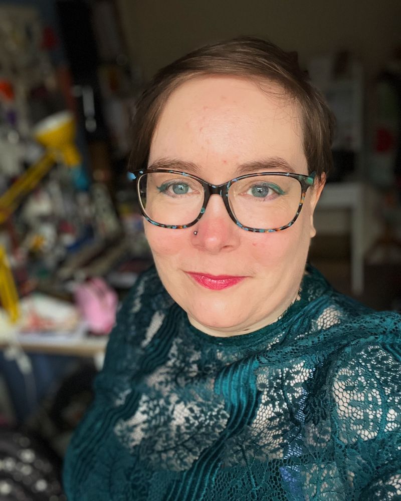 Selfie photo of white nonbinary person with short hair, subtle makeup, glasses and a green lace dress.