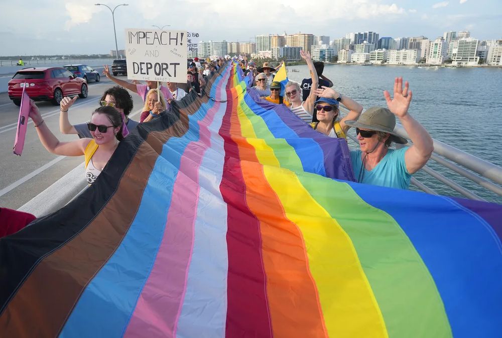No kings protesters, marching across the Ringling Bridge in Sarasota FL, holding a Pride flag spanning the length of the bridge. Downtown Sarasota in the background. 