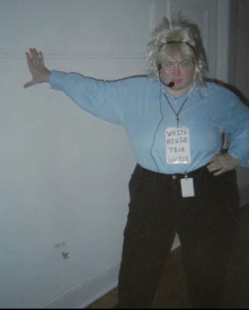 Person in black slacks and a blue shirt and a lanyard that reads “White House tour guide” they are standing with one hand on their hip, their other hand resting on a wall. They are staring directly into the camera. They have glasses and a bleach blonde mullet. 