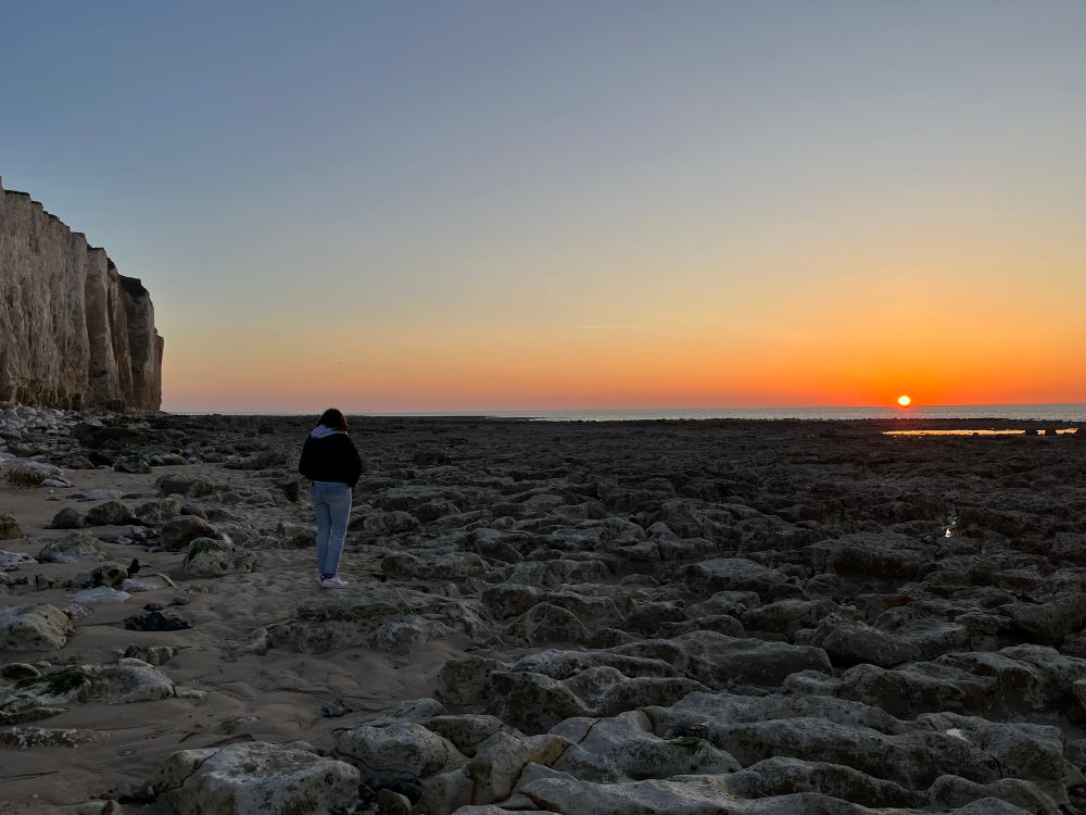 Le soleil se couche , l’horizon est orange , tandis que se détachent à gauche les falaises de craie . Au premier plan , l’estran constitué de blocs crayeux . Une femme, de dos , regarde le coucher de soleil . 