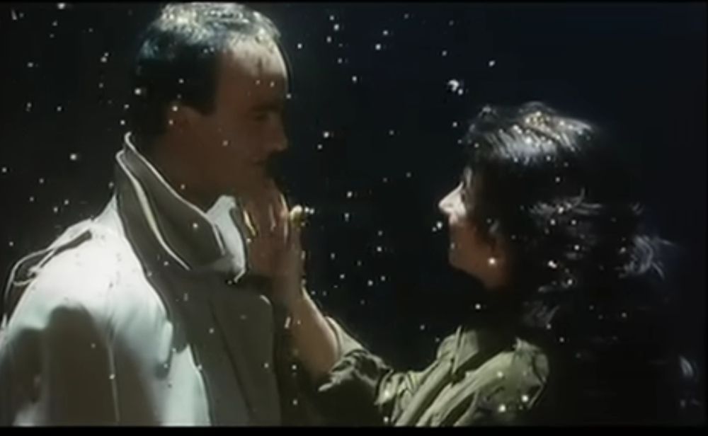 A men in a Trenchcoat looking at a woman, she caresses his cheek 