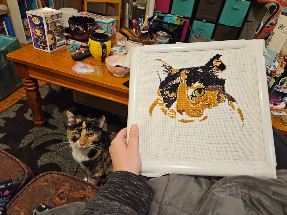 Hannah the tortico cat, sitting on the floor while her mama holds up an unfinished cross-stitch project of a picture of Hannah up next to her. 