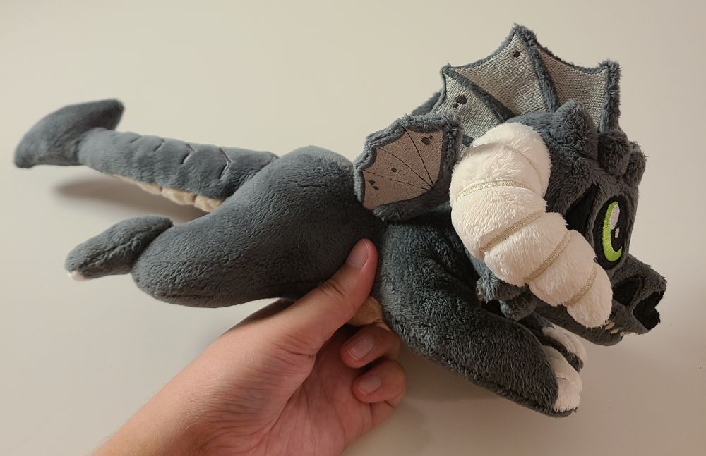 A small, floppy, black dragon plush made by hand. Grey fins, green eyes, cream horns and claws, and skull-like face. Made with soft, minky fabric with embroidered details. Inspired by dungeons and dragons and classic ttrpg games. Filled with light stuffing and polly pellets, the fun textures make this a great plush for fidgeting and stimming.