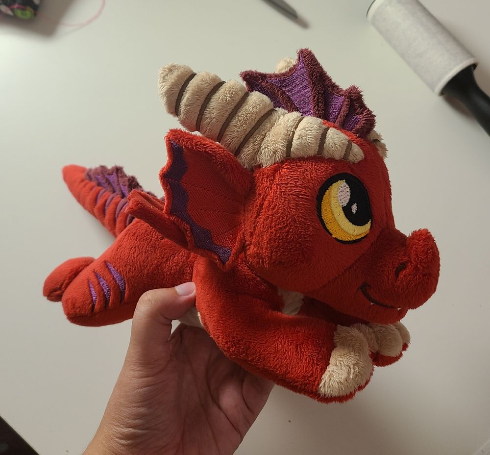 A small, floppy, red dragon plush made by hand. Purple fins, yellow-orange eyes, tan horns and claws, and purple stripes. Made with soft, minky fabric with embroidered details. Inspired by dungeons and dragons and classic ttrpg games. Filled with light stuffing and polly pellets, the fun textures make this a great plush for fidgeting and stimming. For sale!