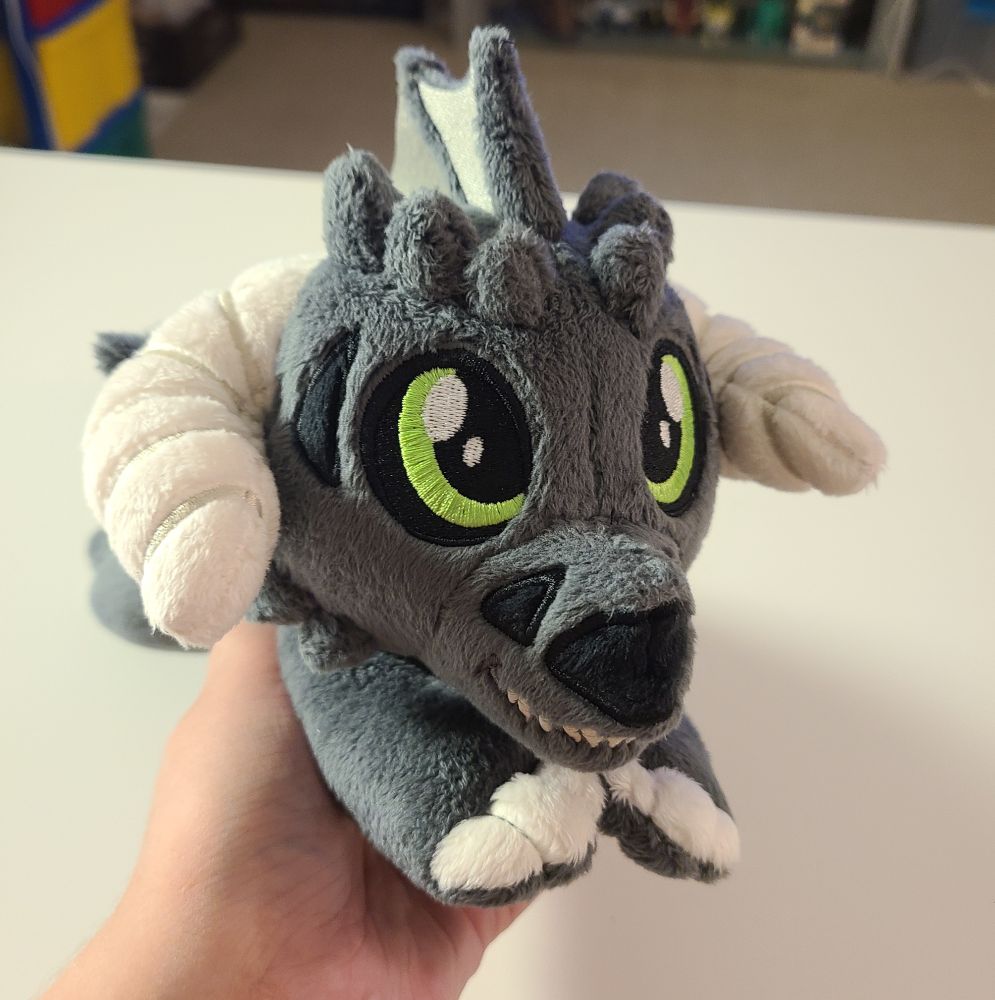 A small, floppy, black dragon plush made by hand. Grey fins, green eyes, cream horns and claws, and skull-like face. Made with soft, minky fabric with embroidered details. Inspired by dungeons and dragons and classic ttrpg games. Filled with light stuffing and polly pellets, the fun textures make this a great plush for fidgeting and stimming.
