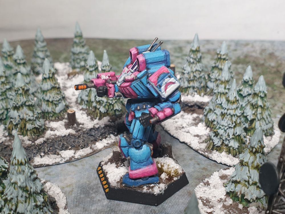 An 8mm scale Battletech mech (a Wolverine, I think) advancing through a snow capped alpine forest, weapon drawn and glowing hot.
The model is painted in cool blues and hot pinks, reminiscent of the Trans Pride flag, and bears a freehanded Blahaj, the Ikea shark, on its left shoulder guard.