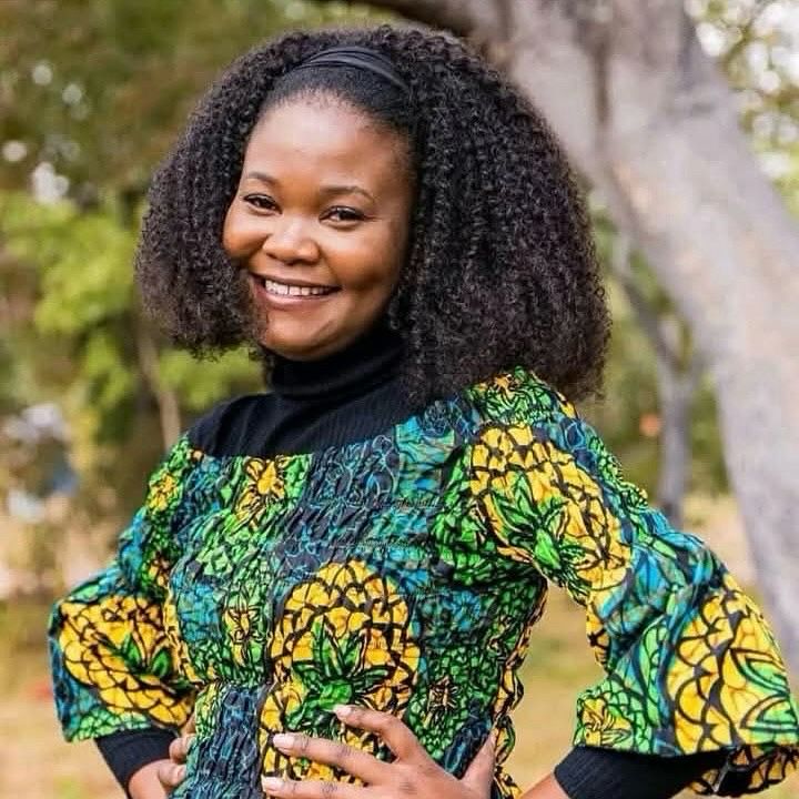 Vibrant photo of Dora wearing a yellow, blue, and green top over a black blouse. She stands outside by a tree with her hands on her hips, smiling cheerfully at the camera.