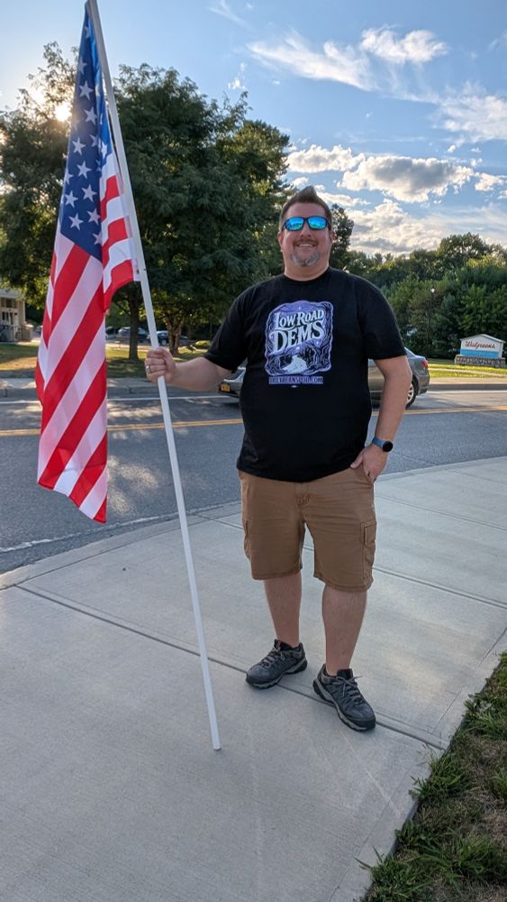 Man holding American Flag at ANTI MAGA Protest. Wearing a tee shirt that says 