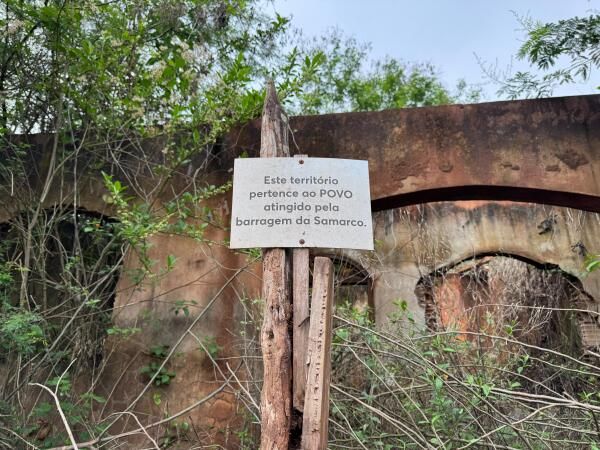 foto em dois planos, no plano de fundo há ruínas de uma construção, rodas as paredes são tingidas de um tom terroso de barro. Há folhagens crescendo junto às ruínas. No plano mais a frente e centralizado, há alguns tocos de madeira com uma placa empoeirada. Essa placa diz "Este território pertence ao POVO atingido pela barragem da Samarco"