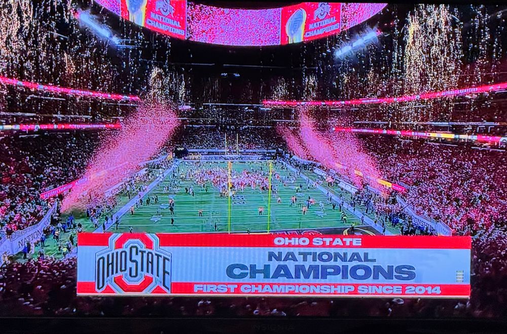 Photo depicts a TV screen with a banner that says “Ohio State - National Champions” while on the football field confetti is raining down and fireworks are going off. 
