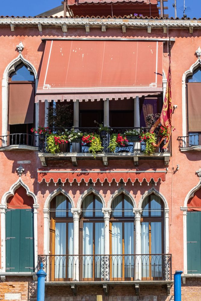 Two floors on the ochre facade of a Venetian palace.
At the level of each floor, a balcony with four high and stylized windows, as well as a canvas blind. 
On the top balcony, flowers in planters and the blind lowered.
On both sides of the balconies, other tall and stylized windows. 
📸: Leandro Silva.