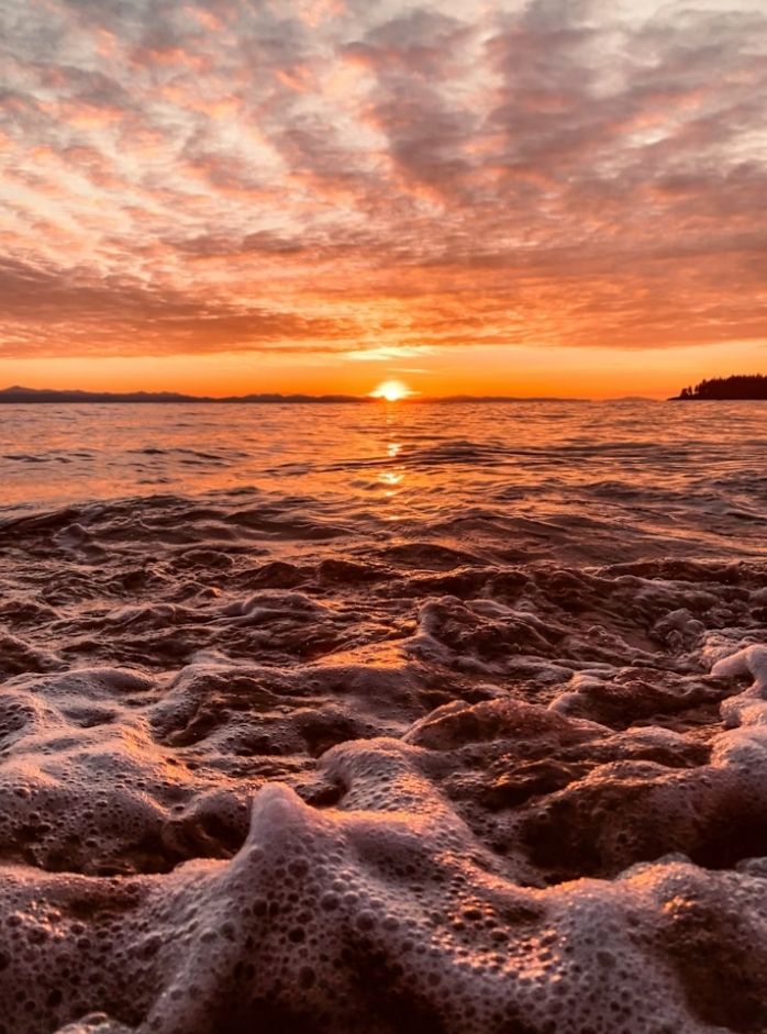A sunset over a rough sea with a close-up of waves and foam. 
The sky is very cloudy, orange, yellow and gray, and is reflected in the water. 
📸: Abhigna Parvath.
