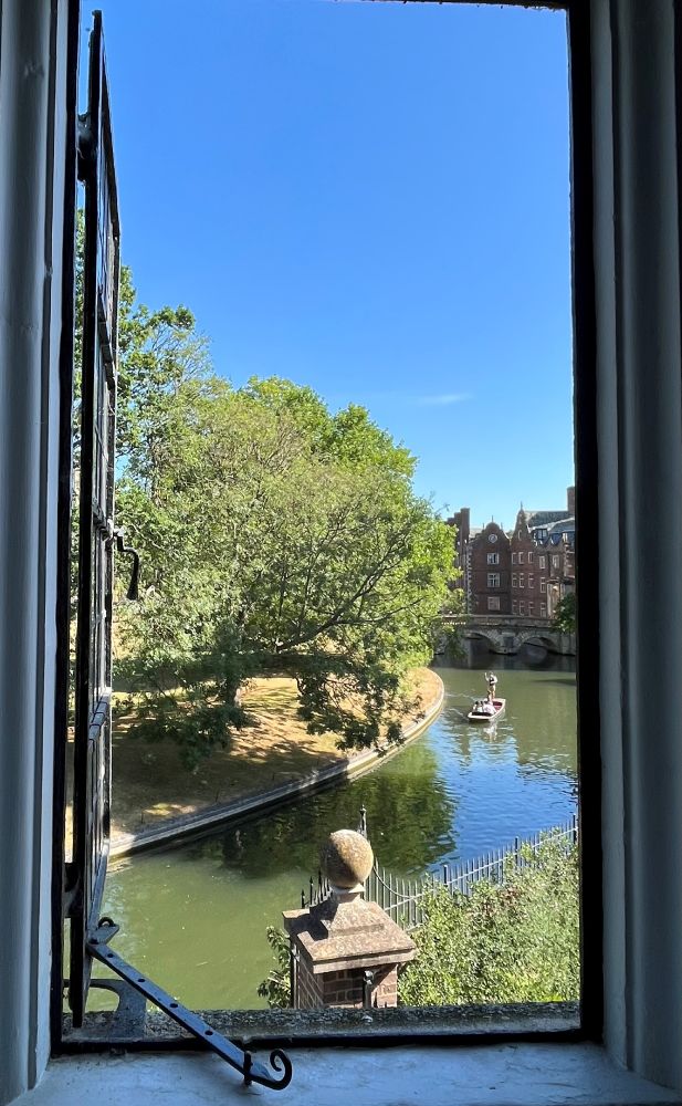 The window view from outside the Wren Library, looking down upon a curving river with a grassy bank and overhanging tree. A punting boat that has just travelled under a bridge appear in the distance.