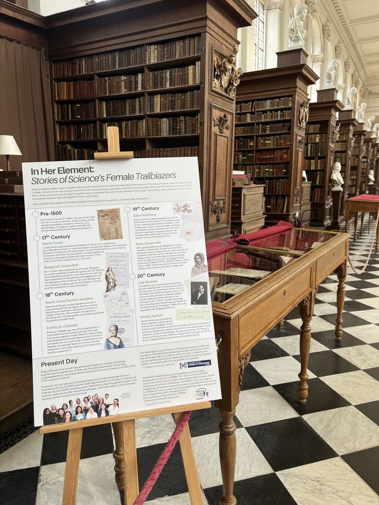 A poster detailing the timeline of the exhibition: 'In Her Element: Stories of Science's Female Trailblazers' is in the foreground. The background includes bays of books in the Wren and open exhibition cases with items inside.