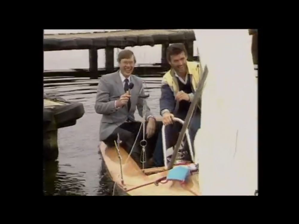 ITV sport commentator, John Helm, sits laughing in a sailing dinghy after his right leg went into the water as he attempted to get into the dinghy to conduct an interview. The other man looks like, but isn’t, Marti Pellow.