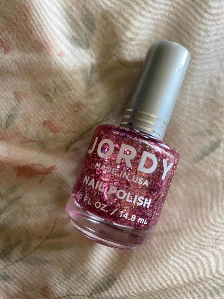 bottle of glittery pink jordy brand nail polish lying flat on floral fabric