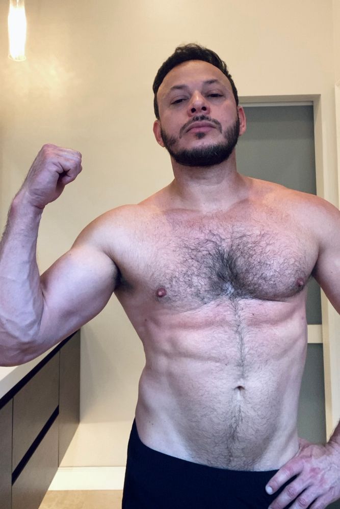 A shirtless man flexes his biceps, showing a hairy chest and slightly visible abs.