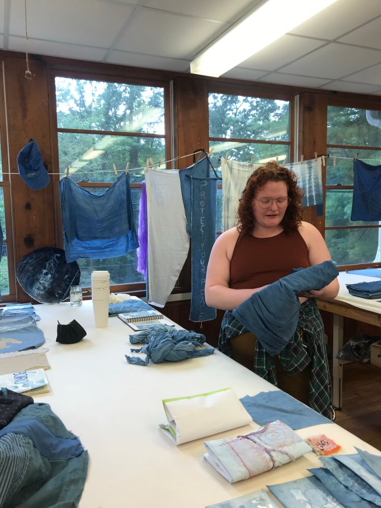 Katie is pictured at their table in the fabric workshop at Penland. They are holding an indigo dyed folded up pair of overalls they made during their visit and have their mouth open in mid-presentation. The table holds a display of various indigo creations, notably a stack of the coffee cups and lids they collected over the course of their two week stay. Windows in the foreground are lined with more indigo dyed projects they began. Katie is wearing their summer uniform, mustard shorts and a deep red tank top with a forrest green, navy blue, and white flannel tied at their waste.