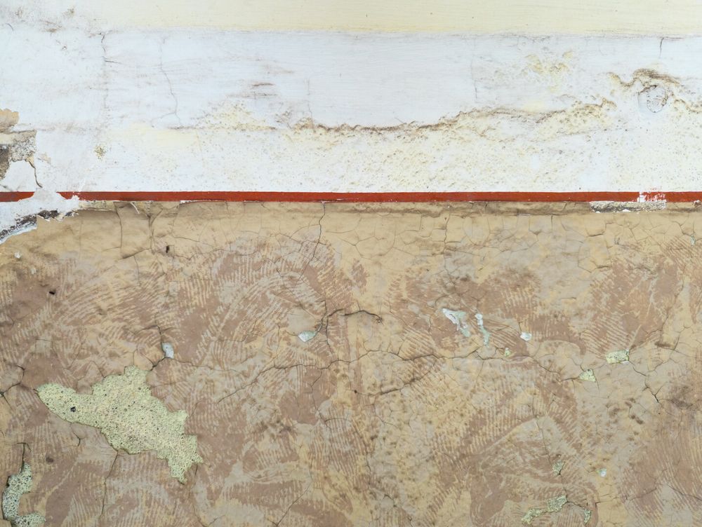 A highly textured and weathered wall is divided horizontally. The top half is off-white with fine cracks, while the bottom half is a darker beige, featuring a network of deep cracks and chipped areas that reveal a different colored layer underneath.