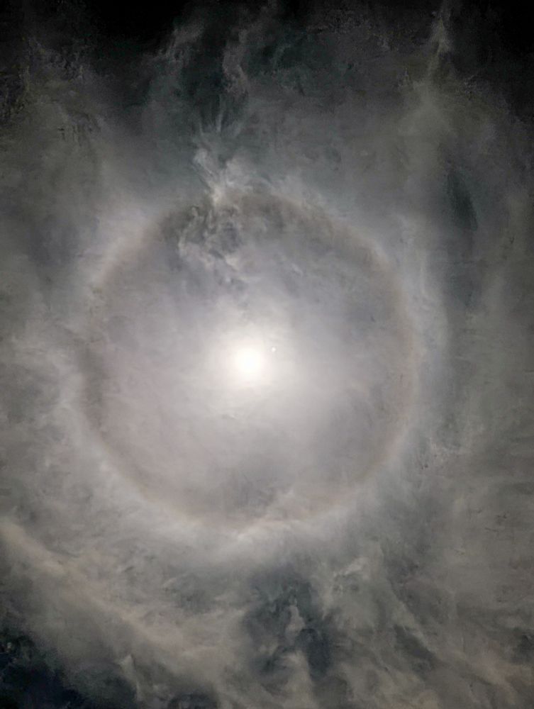 Wispy clouds shroud a bright moon. A hazy white circle can also be seen around the moon.