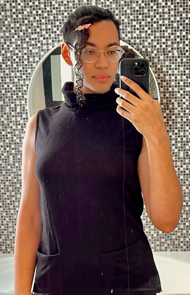 bathroom selfie. i'm wearing a black turtleneck dress with my hair pulled back in a tight bun and a shoulder length strand of bangs falling over the left side of my face. i'm wearing gold rimmed glasses and a magical girl hair pin.