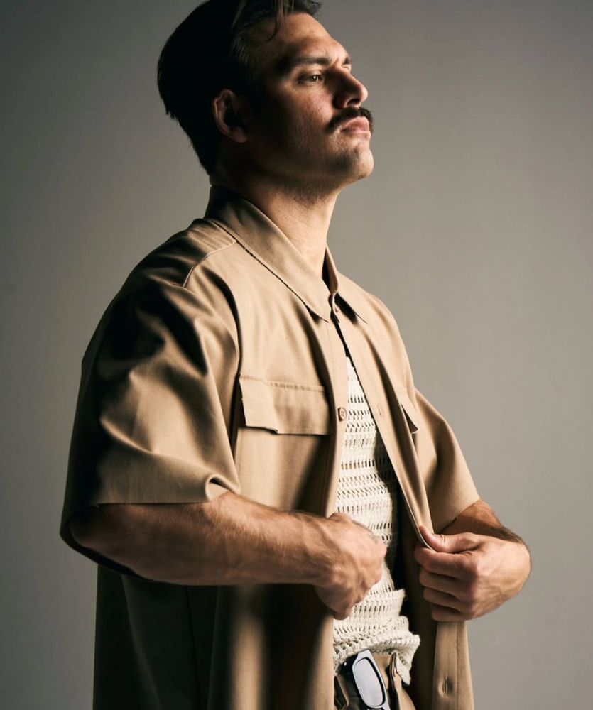 ryan guzman turned mostly to the side. his chin is tilted up a little bit almost like he's posturing. he's wearing a tan short sleeve button up, but it's unbuttoned over a white crocheted shirt of some kind. he's got the sexy mustache, and a serious look on his face