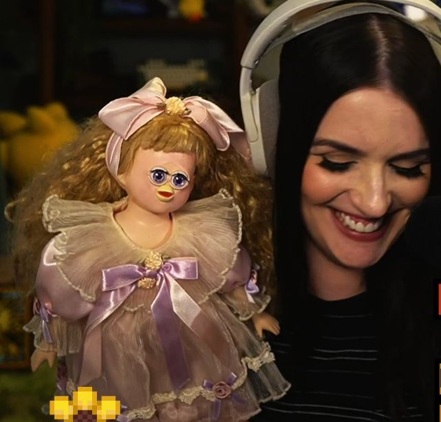 A dark haired woman wearing a headset is smiling while holding a doll that has curly blonde hair, a pink bow, and a frilly pink dress with purple ribbons. It's face is that of a furby with large purple eyes, a bird beak and uncanny features.
