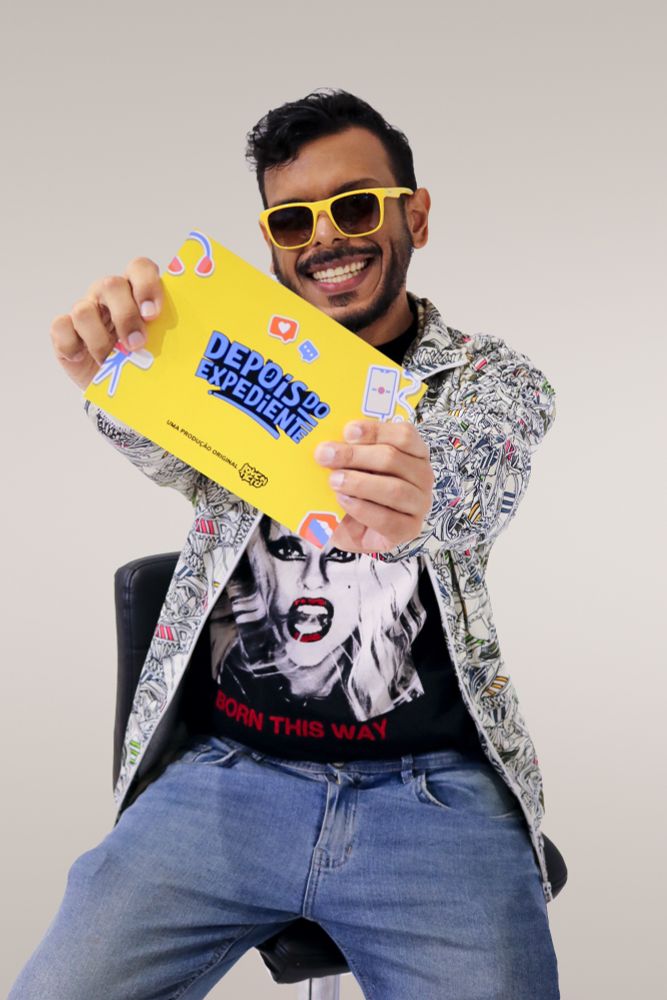 Foto onde estou sentado de jaqueta branca com estampas de tênis, camiseta da Lady Gaga e Jean segurando a ficha de apresentação do Depois do Expediente. Uso um óculos amarelo escuros