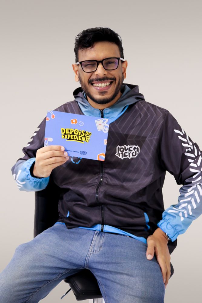 Foto onde estou sentado de jaqueta do Jovem Nerd e jeans segurando a ficha de apresentação do Depois do Expediente. Uso um óculos amarelo escuros
